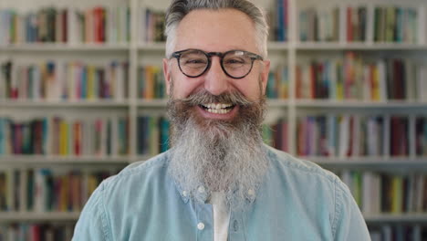 Porträt-Eines-Reifen-Kaukasischen-Professors-Mit-Bart,-Der-Fröhlich-In-Die-Kamera-Im-Bücherregal-Der-Bibliothek-Lacht-Und-Eine-Brille-Trägt