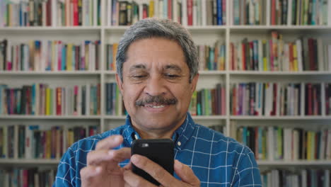 Nahaufnahme-Eines-Porträts-Eines-Glücklichen,-Reifen-Hispanischen-Mannes,-Der-SMS-Schreibt-Und-Mit-Mobiler-Smartphone-Technologie-Surft-Und-Digitale-Kommunikation-Im-Hintergrund-Eines-Bibliotheksbücherregals-Genießt