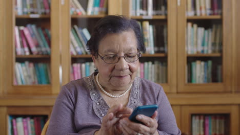 Retrato-De-Una-Anciana-Elegante-En-La-Biblioteca-Escribiendo-Mensajes-De-Texto-Usando-Una-Aplicación-De-Mensajería-Para-Teléfonos-Inteligentes-Sonriendo-Feliz-Con-Gafas