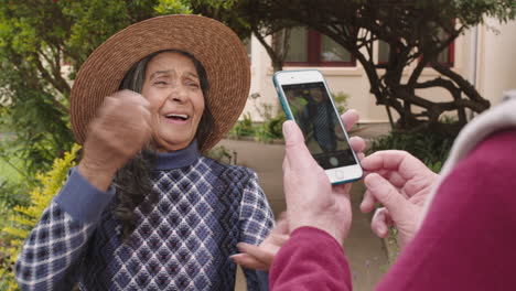 portrait-of-happy-elderly-woman-dancing-posing-wearing-hat-enjoying-retirement-friend-taking-photo-using-smartphone