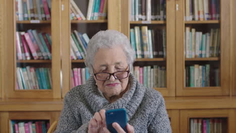 Porträt-Einer-älteren-Frau-In-Der-Bibliothek,-Die-Mit-Der-Smartphone-Messaging-App-SMS-Schreibt-Und-Glücklich-Lächelt-Und-Eine-Brille-Trägt