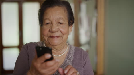 Retrato-De-Una-Anciana-Usando-Un-Teléfono-Inteligente-Disfrutando-Leyendo-Mensajes-En-Línea-Mujer-Mayor-De-Raza-Mixta-Viendo-Videos-En-Un-Teléfono-Móvil-Riendo-Feliz-En-Una-Casa-De-Retiro