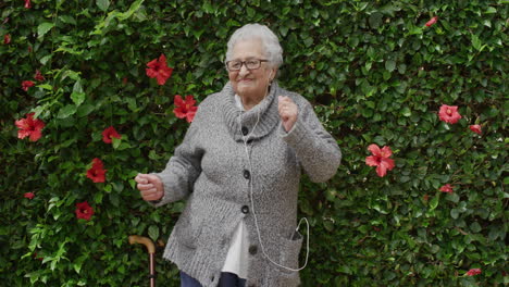 Retrato-De-Una-Alegre-Anciana-Bailando-Usando-Audífonos-Escuchando-Música-Disfrutando-De-Diversión-En-Un-Hermoso-Jardín-Verde-Con-Flores-Al-Aire-Libre-Sosteniendo-Un-Bastón