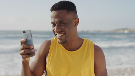 Junger,-Fitter-Afroamerikanischer-Mann,-Der-Lächelnd-Mit-Dem-Smartphone-Fotografiert-Und-Im-Strandurlaub-Mobile-Technologie-Genießt-Und-Eine-Gelbe-Weste-Trägt