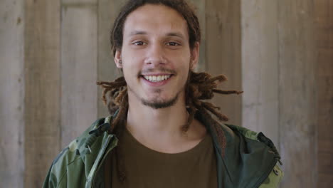 close-up-portrait-of-young-attractive-mixed-race-man-dreadlocks-hairstyle-smiling-looking-at-camera-confident-male-wearing-camouflage-jacket-wooden-background