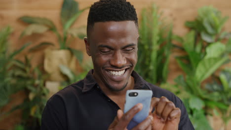 Porträt-Eines-Afroamerikanischen-Mannes,-Der-Sein-Smartphone-Nutzt-Und-Lächelnd-Online-Nachrichten-Durchstöbert-Und-Dabei-Genießt,-In-Zeitlupe-SMS-Zu-Schreiben