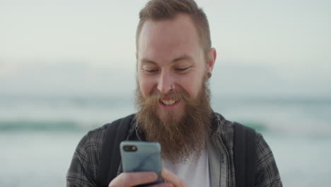 Porträt-Eines-Jungen-Hipster-Mannes,-Der-Lächelnd-Sein-Smartphone-Nutzt-Und-Dabei-Genießt,-SMS-Zu-Schreiben,-Im-Internet-Zu-Surfen,-Am-Strand-Zu-Surfen-Und-Urlaubserlebnisse-Zu-Teilen