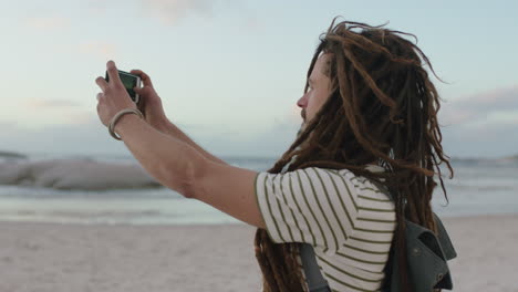 young-carefree-man-with-dreadlocks-taking-photos-on-beach-using-phone-wearing-stripe-shirt