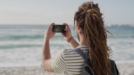 Junger-Touristenmann,-Der-Mit-Smartphone-Kameratechnologie-Am-Strand-Ein-Foto-Von-Einem-Wunderschönen-Malerischen-Ozeanhintergrund-Macht,-Kaukasischer-Mann,-Der-Die-Sommerferienreise-Genießt