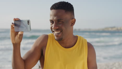 Junger-Gutaussehender-Afroamerikanischer-Mann,-Der-Lächelnd-Mit-Dem-Smartphone-Fotografiert-Und-Im-Strandurlaub-Mobile-Technologie-Genießt-Und-Eine-Gelbe-Weste-Trägt