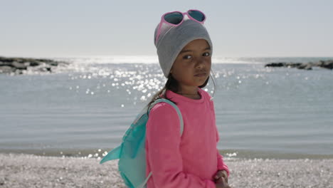 portrait-of-little-girl-on-beach-turning-looking-wearing-backpack-beanie-pink-clothes