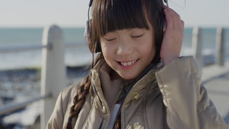 Nahaufnahme-Eines-Porträts-Eines-Niedlichen-Kleinen-Asiatischen-Mädchens-Mit-Kopfhörern,-Das-Lächelnd-Tanzt-Und-Das-Musikhören-Am-Strand-Genießt
