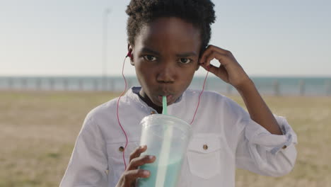 Primer-Plano-Retrato-De-Un-Animado-Niño-Afroamericano-Disfrutando-Bebiendo-Batido-Usando-Auriculares-Escuchando-Música-Bailando-Juguetón-En-El-Parque