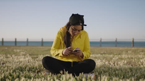 portrait-of-mixed-race-teenage-girl-sitting-at-seaside-park-texting-browsing-using-smarphone-social-media-app-enjoying-relaxed