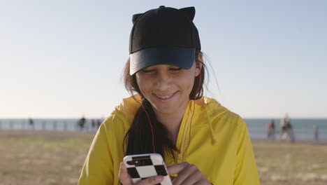Nahaufnahme-Eines-Porträts-Eines-Teenager-Mädchens,-Das-SMS-Schreibt-Und-Mit-Dem-Smartphone-Surft,-Kopfhörer-Trägt-Und-Im-Park-Am-Meer-Musik-Hört-Und-Einen-Süßen-Hut-Trägt