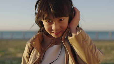 Nahaufnahme-Eines-Süßen-Kleinen-Asiatischen-Mädchens-Mit-Kopfhörern,-Das-Lächelt-Und-Bei-Sonnenuntergang-Musik-Im-Strandpark-Am-Meer-Hört