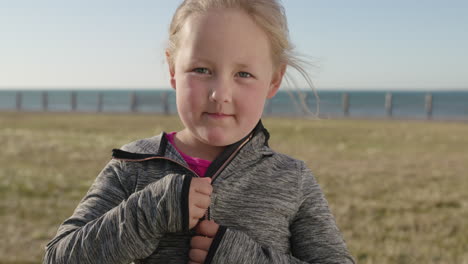 close-up-portrait-of-little-blonde-girl-smiling-at-camera-comfortable-wearing-cozy-hoodie-enjoying-sunny-seaside-park
