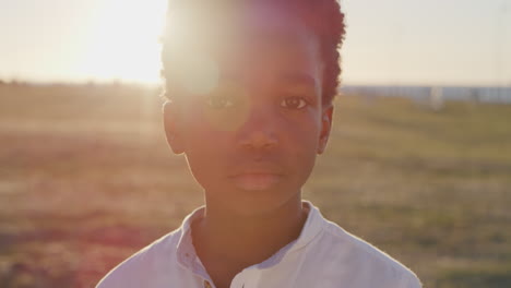 Nahaufnahme-Eines-Porträts-Eines-Kleinen-Afroamerikanischen-Jungen,-Der-Ernst-Und-Nachdenklich-In-Die-Kamera-Blickt,-Auf-Dem-Hintergrund-Des-Sonnenuntergangs-Im-Strandpark-Am-Meer-Mit-Blendenfleck
