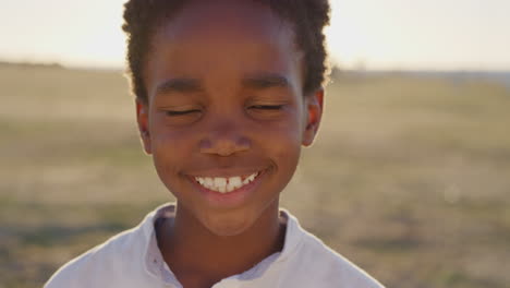 Nahaufnahme-Eines-Porträts-Eines-Süßen-Afroamerikanischen-Jungen,-Der-Fröhlich-Lächelt-Und-In-Die-Kamera-Blickt-Und-Glücklich-Den-Sonnigen-Tag-Im-Park-Am-Meer-Bei-Hellem,-Lebendigem-Sonnenuntergang-Genießt