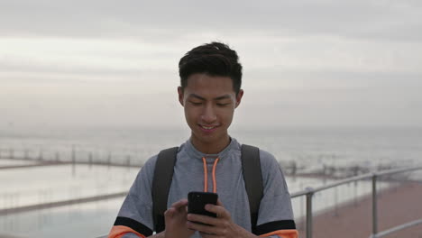 Porträt-Eines-Jungen-Asiatischen-Mannes,-Der-Am-Bewölkten-Meer-Eine-SMS-Schreibt-Und-Mit-Dem-Telefon-Surft