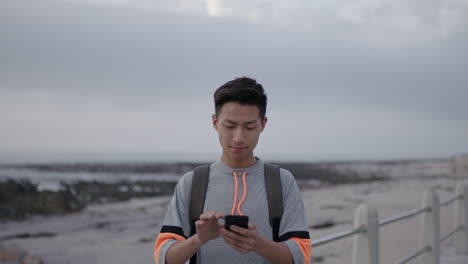 Porträt-Eines-Jungen-Asiatischen-Mannes,-Der-Am-Strand-Am-Meer-Steht-Und-Per-Telefon-SMS-Schreibt