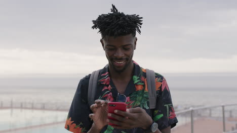 Porträt-Eines-Jungen-Afroamerikanischen-Mannes,-Der-SMS-Schreibt-Und-Mit-Dem-Smartphone-Surft-Und-Lächelt-Und-Einen-Glücklichen-Urlaub-Am-Meer-Genießt