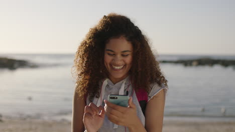 portrait-of-mixed-raced-woman-with-frizzy-hair-enjoying-sunny-day-on-beach-smiling-using-phone-texting