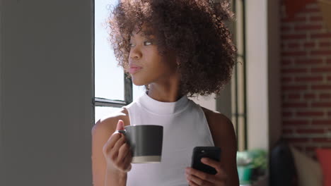 Hermosa-Mujer-Usando-Un-Teléfono-Inteligente-Tomando-Café-En-Casa-Disfrutando-De-Una-Mañana-Relajada-Navegando-Mensajes-Mirando-Por-La-Ventana-Planificando-Con-Anticipación-Una-Mujer-Negra-Con-Un-Moderno-Peinado-Afro