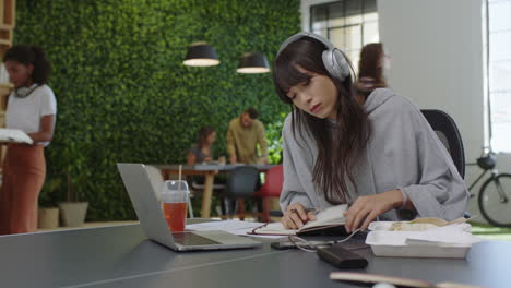 young-asian-business-woman-writing-notes-enjoying-study-listening-to-music-wearing-headphones-female-student-using-smartphone-checking-messages-in-diverse-office-workplace