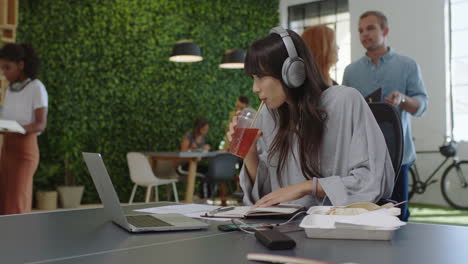 young-asian-business-woman-writing-notes-enjoying-study-listening-to-music-wearing-headphones-female-student-drinking-juice-in-diverse-office-workplace