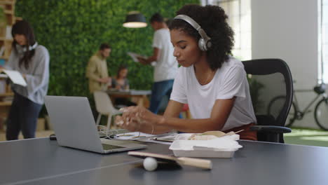 Junge-Afroamerikanische-Geschäftsfrau-Schreibt-Notizen-Und-Genießt-Das-Studium,-Hört-Musik.-Schwarze-Studentin-Nutzt-Laptop-Und-Recherchiert-Online-An-Einem-Geschäftigen-Büroarbeitsplatz