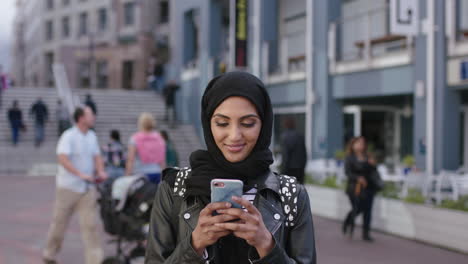 Retrato-De-Una-Joven-Mujer-Del-Medio-Oriente-Mirando-Un-Teléfono-Inteligente-Sorprendida-Emocionada-En-El-Fondo-Urbano