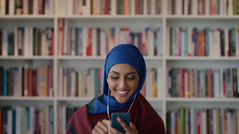 Retrato-Hermosa-Joven-Estudiante-Musulmana-Usando-Teléfonos-Inteligentes-Enviando-Mensajes-De-Texto-Navegando-En-Línea-Disfrutando-Escuchando-Música-Usando-Hijab-En-El-Fondo-De-La-Estantería-De-La-Biblioteca-En-Cámara-Lenta