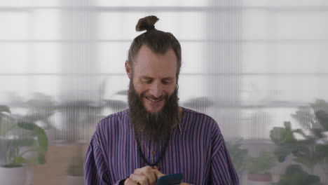 portrait-of-mature-hippie-entrepreneur-man-smiling-enjoying-texting-browsing-using-smartphone-mobile-technology-businessman-with-beard-in-office-background