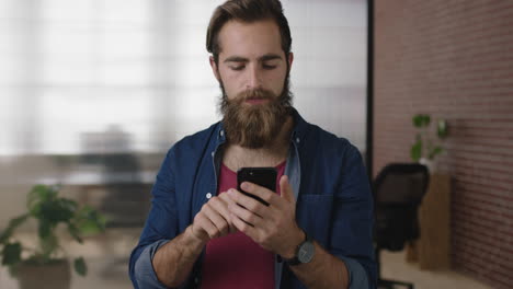 portrait-of-attractive-young-hipster-man-with-beard-texting-browsing-checking-messages-using-smartphone-mobile-app-in-office-background