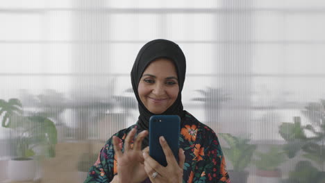 Retrato-De-Una-Mujer-Musulmana-Mayor-Disfrutando-De-Mensajes-De-Texto-Navegando-En-Línea-Usando-Un-Teléfono-Inteligente-Sonriendo-Feliz-Mujer-De-Mediana-Edad-En-El-Fondo-De-La-Oficina