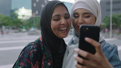 Retrato-De-Cerca-De-Madre-E-Hija-Musulmanas-Sonriendo-Alegremente-Haciendo-Caras-Posando-Tomando-Una-Foto-Selfie-Usando-Un-Teléfono-Inteligente-En-Una-Ciudad-Urbana