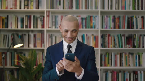 close-up-portrait-of--bald-hispanic-businessman-using-smartphone-smiling-texting-social-media-bookshelf-library