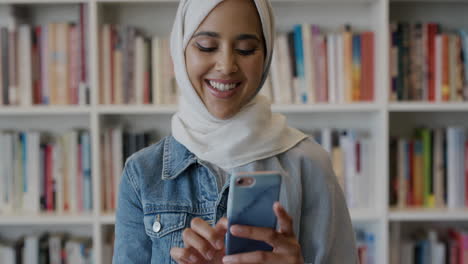 Porträt-Einer-Jungen,-Glücklichen-Muslimischen-Studentin,-Die-Ihr-Smartphone-Nutzt-Und-Spaß-Daran-Hat,-Online-Zu-Surfen-Und-Nachrichten-über-Die-Kommunikations-App-Für-Mobiltelefone-Zu-Senden,-Die-Ein-Hijab-Kopftuch-Im-Hintergrund-Eines-Bibliotheksbücherregals-Trägt