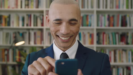Nahaufnahme-Porträt-Eines-Hispanischen-Geschäftsmannes-Mit-Glatze,-Der-Sein-Smartphone-Lächelt-Und-SMS-In-Der-Bücherregalbibliothek-In-Den-Sozialen-Medien-Schreibt