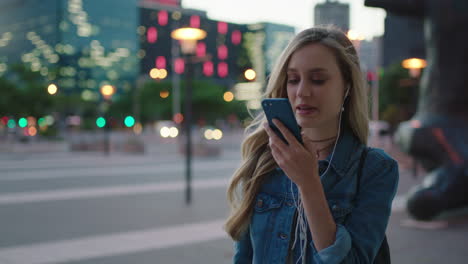 portrait-of-attractive-blonde-girl-wearing-earphones-talking-on-smartphone-to-friend-enjoying-calm-urban-evening-outdoors-in-city