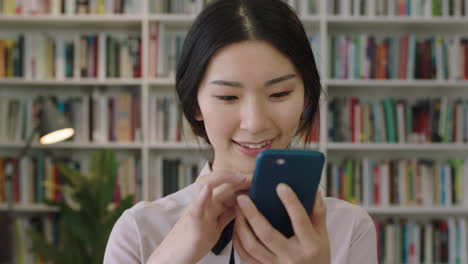 Nahaufnahme-Porträt-Einer-Schönen,-Süßen-Asiatischen-Frau,-Die-In-Der-Bibliothek-Steht-Und-Ihr-Smartphone-Benutzt,-Lächelnd-Und-Lachend-Im-Bücherregal-Im-Hintergrund