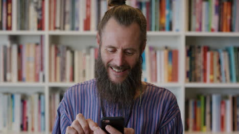 portrait-mature-hippie-entrepreneur-man-using-smartphone-enjoying-browsing-online-messages-sending-email-communication-on-mobile-phone-bearded-caucasian-male-in-bookshelf-background