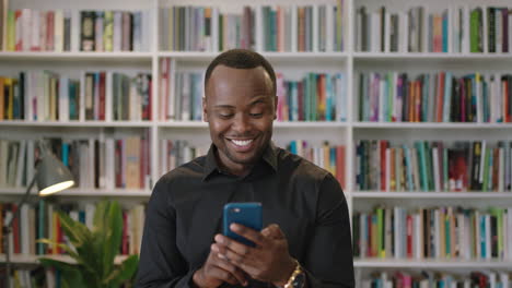 Porträt-Eines-Jungen-Afroamerikanischen-Mannes-Mit-Smartphone,-Der-Lachend-In-Den-Sozialen-Medien-Der-Bibliothek-Steht