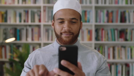 Joven-Hombre-Confiado-Del-Medio-Oriente-Parado-En-La-Biblioteca-Usando-Un-Teléfono-Inteligente-Retrato-Sonriente-De-Un-Orgulloso-Empresario-Usando-Las-Redes-Sociales