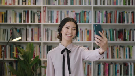 Porträt-Einer-Schönen-Asiatischen-Frau,-Die-In-Einer-Bibliothek-Steht-Und-Mit-Ihrem-Smartphone-Ein-Video-Chattet,-Ein-Selfie-Bücherregal-Im-Hintergrund,-Lächelnd-Und-Winkend