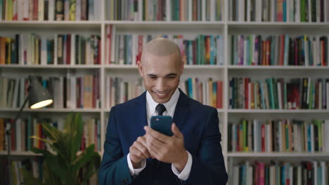 Nahaufnahme-Eines-Kahlköpfigen-Hispanischen-Geschäftsmannes,-Der-Sein-Smartphone-Lächelt-Und-SMS-In-Der-Bücherregalbibliothek-In-Den-Sozialen-Medien-Schreibt