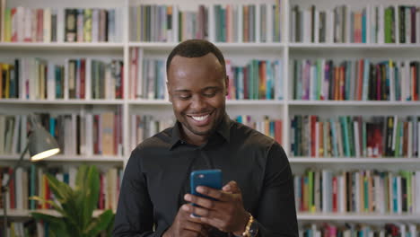 Porträt-Eines-Jungen-Afroamerikanischen-Mannes-Mit-Smartphone,-Der-Lachend-In-Den-Sozialen-Medien-Der-Bibliothek-Steht