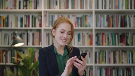 portrait-of-beautiful-young-red-head-business-woman-executive-smiling-enjoying-texting-browsing-using-smartphone-mobile-app-in-library-background