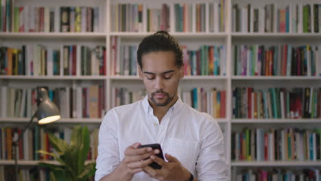 portrait-of-young-hispanic-man-student-enjoying-texting-browsing-online-using-smartphone-mobile-app-looking-at-camera-in-library-bookshelf-background